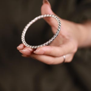 May include: A silver bangle with a twisted rope pattern. The bracelet is held in a hand with manicured nails. The background is a blurred, dark green colour.