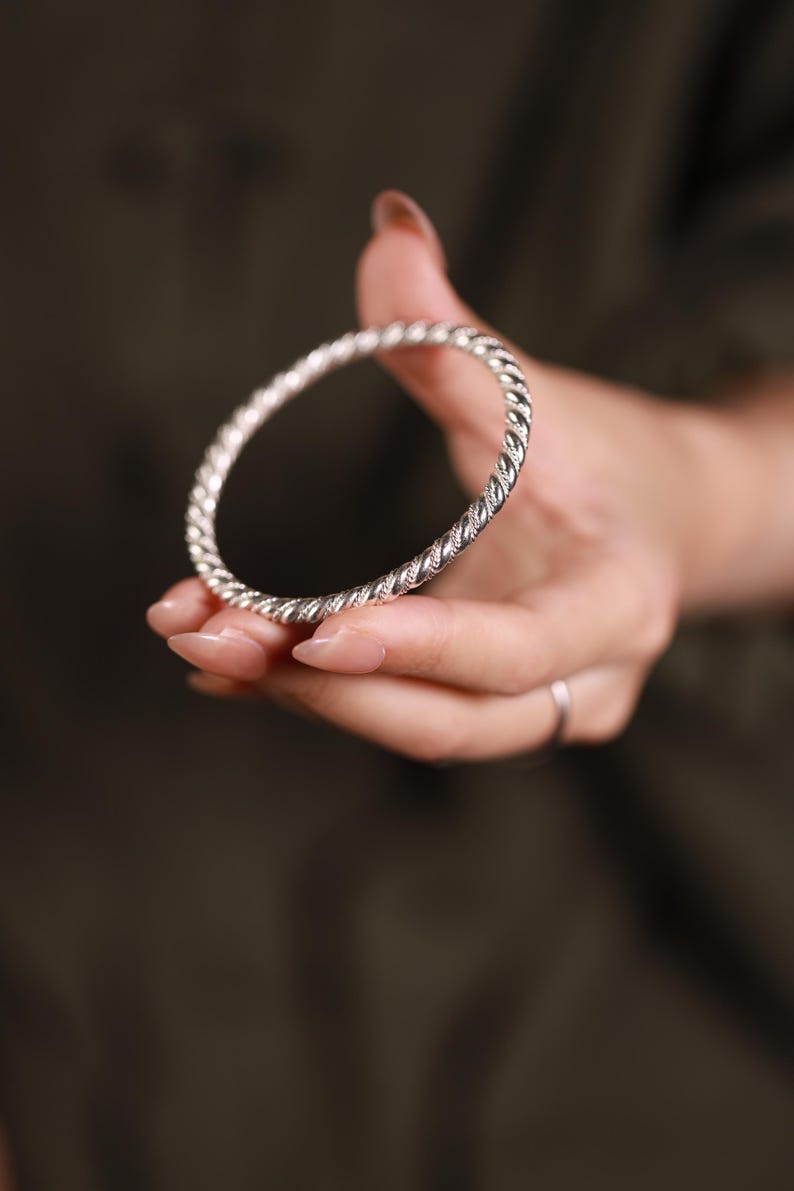 May include: A silver bangle with a twisted rope pattern. The bracelet is held in a hand with manicured nails. The background is a blurred, dark green colour.