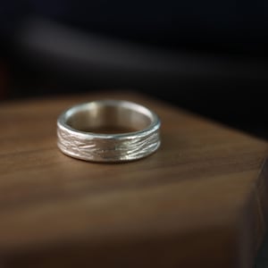 Könnte beinhalten: Ein silberner Ring mit strukturierter Oberfläche, der auf einem Holzblock liegt. Das Band des Rings hat ein detailliertes, organisches Muster. Der Ring ist ein Schmuckstück.