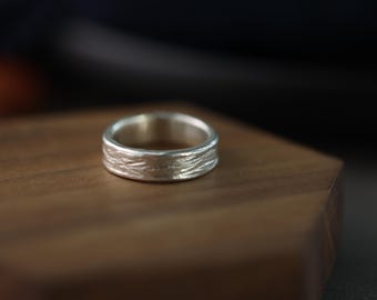 May include: A silver ring with a textured surface, resting on a wooden block. The ring's band has a detailed, organic pattern. The ring is a piece of jewelry.
