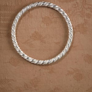 May include: A silver bangle with a twisted rope pattern. The circular bracelet is made of metal. The background is a textured brown surface. The bangle's design is intricate.