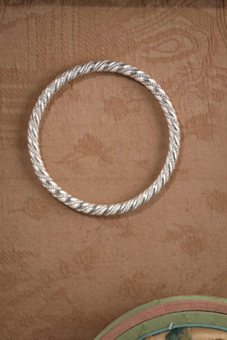 May include: A silver bangle with a twisted rope pattern. The circular bracelet is made of metal. The background is a textured brown surface. The bangle's design is intricate.