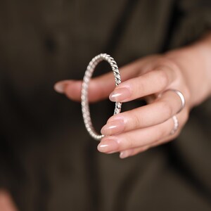 May include: A silver bangle with a twisted rope pattern. The bracelet is held in a hand with long, manicured nails. The background is a dark, solid colour.