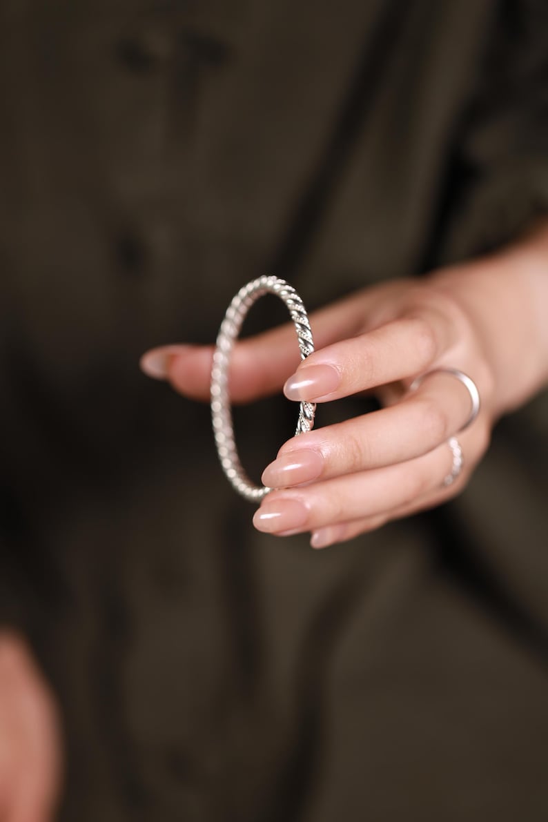 May include: A silver bangle with a twisted rope pattern. The bracelet is held in a hand with long, manicured nails. The background is a dark, solid colour.