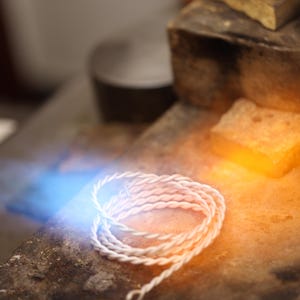 May include: Close-up of a white, twisted wire jewellery piece, possibly a bracelet or necklace component, coiled on a textured surface. The image is illuminated with blue and orange light, suggesting a jeweller's workspace.