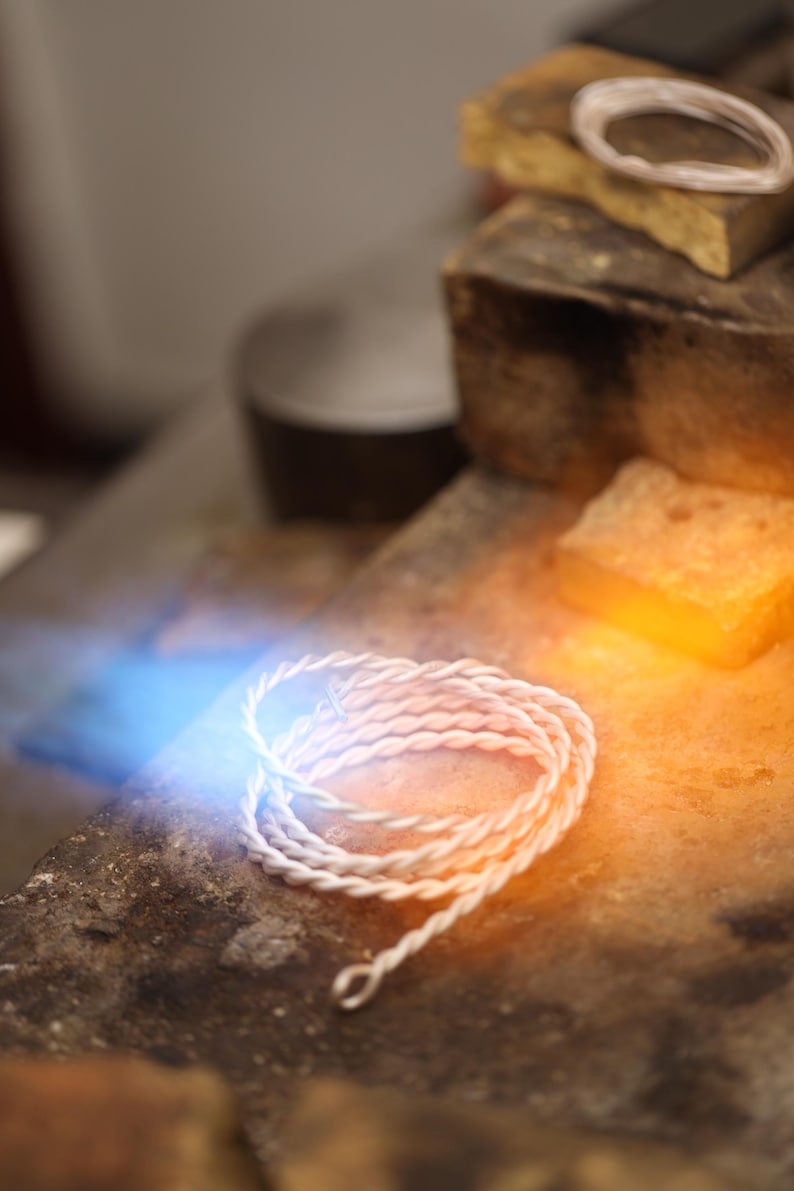 May include: Close-up of a white, twisted wire jewellery piece, possibly a bracelet or necklace component, coiled on a textured surface. The image is illuminated with blue and orange light, suggesting a jeweller's workspace.