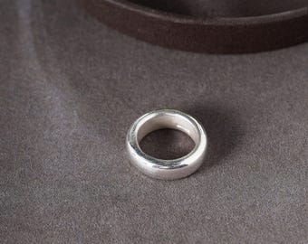 May include: A silver ring with a smooth, rounded design. The ring is made of a shiny, metallic material and sits on a gray surface. The ring's simple, classic style makes it suitable for everyday wear.