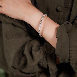May include: A silver-coloured, twisted rope design bracelet. The bracelet is worn on an arm, against a dark green background. The bracelet is a simple, elegant piece of jewellery.