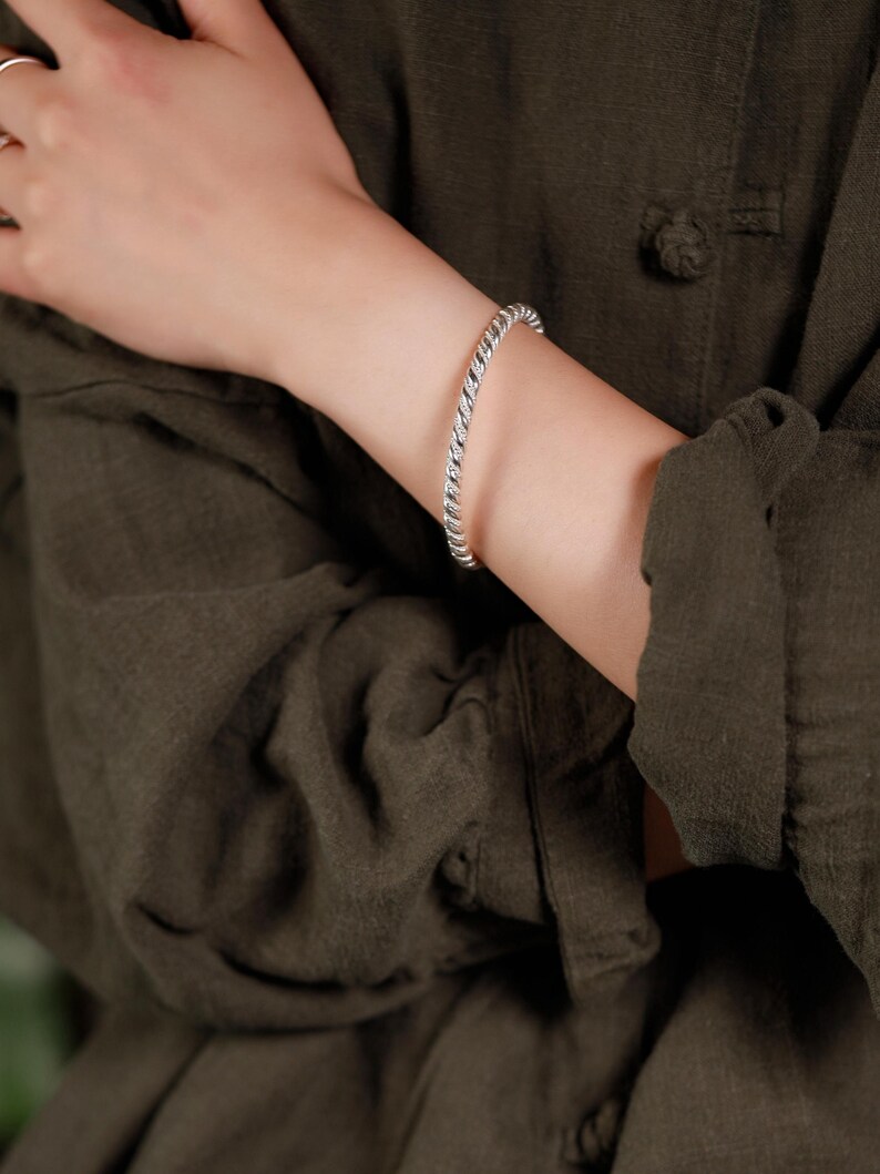 May include: A silver-coloured, twisted rope design bracelet. The bracelet is worn on an arm, against a dark green background. The bracelet is a simple, elegant piece of jewellery.