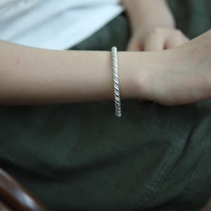 May include: A silver-coloured twisted rope bracelet. The bracelet is worn on a person's wrist. The bracelet's design features a series of tightly wound, textured strands, creating a rope-like appearance. The bracelet is a simple, elegant piece of jewellery.