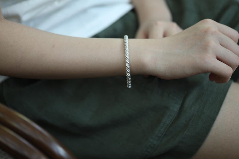 May include: A silver-coloured twisted rope bracelet. The bracelet is worn on a person's wrist. The bracelet's design features a series of tightly wound, textured strands, creating a rope-like appearance. The bracelet is a simple, elegant piece of jewellery.