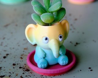 May include: A small elephant-shaped planter featuring a gradient of yellow, light green, and blue colors. The planter holds a green succulent plant and sits on a circular pink saucer. The elephant has large ears, a trunk, and two eyes.