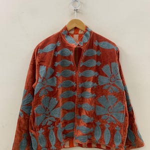 Può includere: Giacca in velluto color ruggine con ricami floreali blu. La giacca presenta un collo alla coreana e maniche lunghe. Il ricamo include grandi fiori e forme più piccole a forma di foglia. La giacca è appesa a una gruccia di legno.