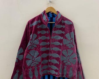 Puede incluir: Chaqueta de terciopelo burdeos con diseños florales y abstractos bordados en gris. La chaqueta tiene un cuello alto y está abierta por delante, revelando un forro a rayas azules y negras. La chaqueta está colgada de una percha de madera.