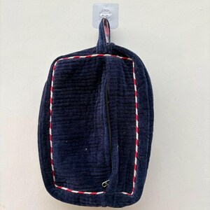 May include: A rectangular navy blue quilted fabric pouch featuring a vertical zippered pocket on the front. The pouch is accented with a red and white twisted cord trim along the edges and includes a fabric loop at the top for hanging.
