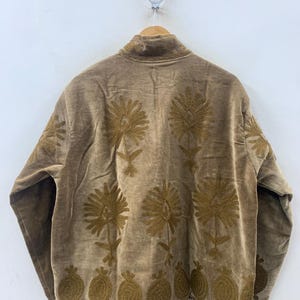 May include: A brown jacket with intricate gold floral embroidery. The jacket features a high collar and is hanging on a wooden hanger. The embroidery includes large flower and leaf designs, as well as a row of pomegranate-like shapes.