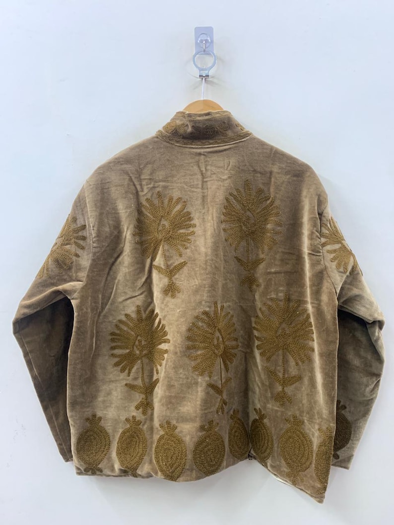 May include: A brown jacket with intricate gold floral embroidery. The jacket features a high collar and is hanging on a wooden hanger. The embroidery includes large flower and leaf designs, as well as a row of pomegranate-like shapes.