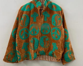 May include: A brown velvet jacket with a green embroidered pattern of peace signs, mushrooms, and yin-yang symbols. The jacket has a striped lining and a collarless design. The jacket is hanging on a wooden hanger.