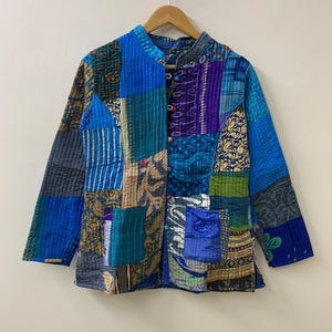 Könnte beinhalten: Eine Patchwork-Jacke in Blau-, Türkis- und Lilatönen mit Stehkragen und Knopfverschluss. Die Jacke besteht aus verschiedenen Stoffmustern und -texturen, wodurch ein einzigartiges und künstlerisches Design entsteht. Sie hängt an einem Holzbügel vor neutralem Hintergrund.