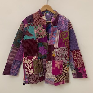 May include: A patchwork jacket in shades of purple, burgundy, and teal, with floral and geometric patterns. The jacket has a mandarin collar and button closure. The jacket is hanging on a wooden hanger.