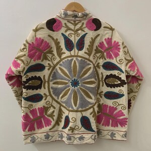 Puede incluir: Chaqueta color crema con intrincados bordados florales en rosa, azul, marr&oacute;n y verde. La chaqueta presenta un cuello alto y un dise&ntilde;o sim&eacute;trico detallado en la espalda. El bordado incluye flores estilizadas y patrones de hojas.