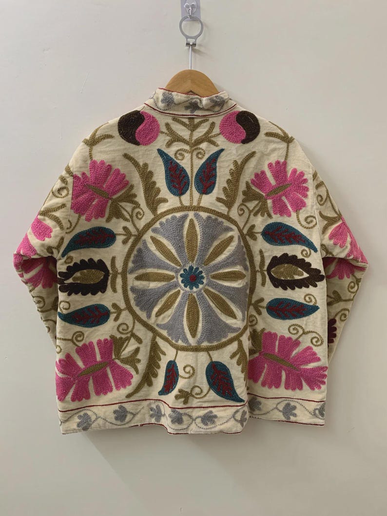 Puede incluir: Chaqueta color crema con intrincados bordados florales en rosa, azul, marr&oacute;n y verde. La chaqueta presenta un cuello alto y un dise&ntilde;o sim&eacute;trico detallado en la espalda. El bordado incluye flores estilizadas y patrones de hojas.