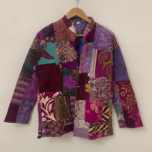 Op de afbeelding: Een patchwork jas in tinten paars, bordeauxrood en blauwgroen, met bloemen- en geometrische patronen. De jas heeft een opstaande kraag en knoopsluitingen. Het kledingstuk hangt aan een houten hanger.