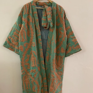 May include: A long, open-front robe in a soft green hue, decorated with an intricate, raised floral pattern in a warm orange-brown. The robe has wide sleeves and a matching tie, with a striped lining visible at the collar and inside. The garment hangs on a wooden hanger.