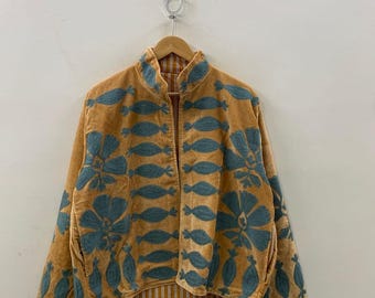 May include: A mustard-colored jacket with teal floral embroidery. The jacket has a high collar and a striped lining. The embroidery features large flowers and smaller, stylized leaf shapes. The jacket is hanging on a wooden hanger.