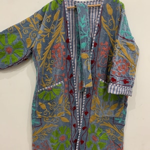 May include: A long, open-front robe in a muted blue hue, with intricate floral embroidery in green, gold, and coral. The robe has two pockets and a striped lining. The garment is hanging on a wooden hanger.