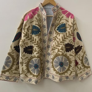 Puede incluir: Chaqueta beige bordada con motivos florales y de hojas en tonos rosa, azul, marr&oacute;n y gris. La chaqueta tiene cuello mandar&iacute;n y est&aacute; forrada con una tela a rayas. Cuelga de una percha de madera.