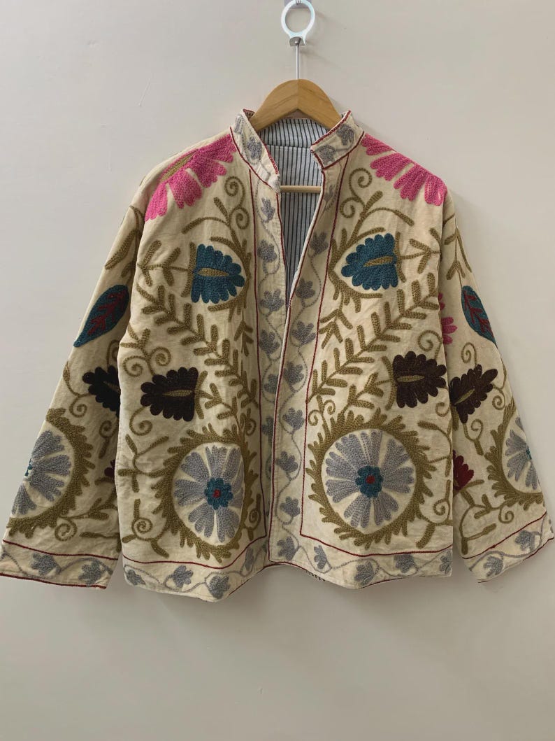 Puede incluir: Chaqueta beige bordada con motivos florales y de hojas en tonos rosa, azul, marr&oacute;n y gris. La chaqueta tiene cuello mandar&iacute;n y est&aacute; forrada con una tela a rayas. Cuelga de una percha de madera.