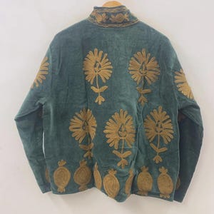 May include: A teal jacket with intricate gold floral embroidery. The jacket features a high collar and long sleeves. The floral design is repeated across the front and back of the jacket, creating a decorative pattern. The jacket is hanging on a wooden hanger.