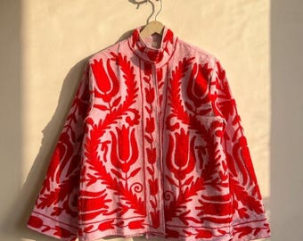 Puede incluir: Chaqueta rosa claro con cuello alto y mangas largas. La chaqueta está adornada con un llamativo diseño de bordado floral rojo. La chaqueta está colgada de una percha de madera.