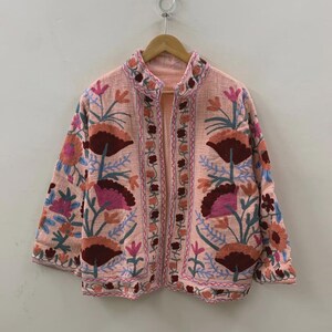May include: A pale pink jacket adorned with detailed floral embroidery in burgundy, pink, and blue hues. The jacket has a mandarin collar and a button-up closure. The floral pattern is repeated on the sleeves and edges of the jacket.