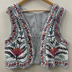 Puede incluir: Chaleco blanco bordado con motivos florales y de hojas en rosa, naranja, verde y negro. El chaleco tiene un forro a rayas y se muestra en una percha de madera. El bordado es detallado y cubre los bordes delanteros y la parte inferior.
