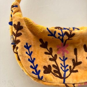 Pu&ograve; includere: Una borsa a tracolla a forma di mezzaluna realizzata in tessuto di velluto giallo. La borsa presenta un intricato ricamo con fili marrone scuro, blu e rosa, che raffigura vari motivi floreali e botanici sulla parte anteriore e sulla tracolla.