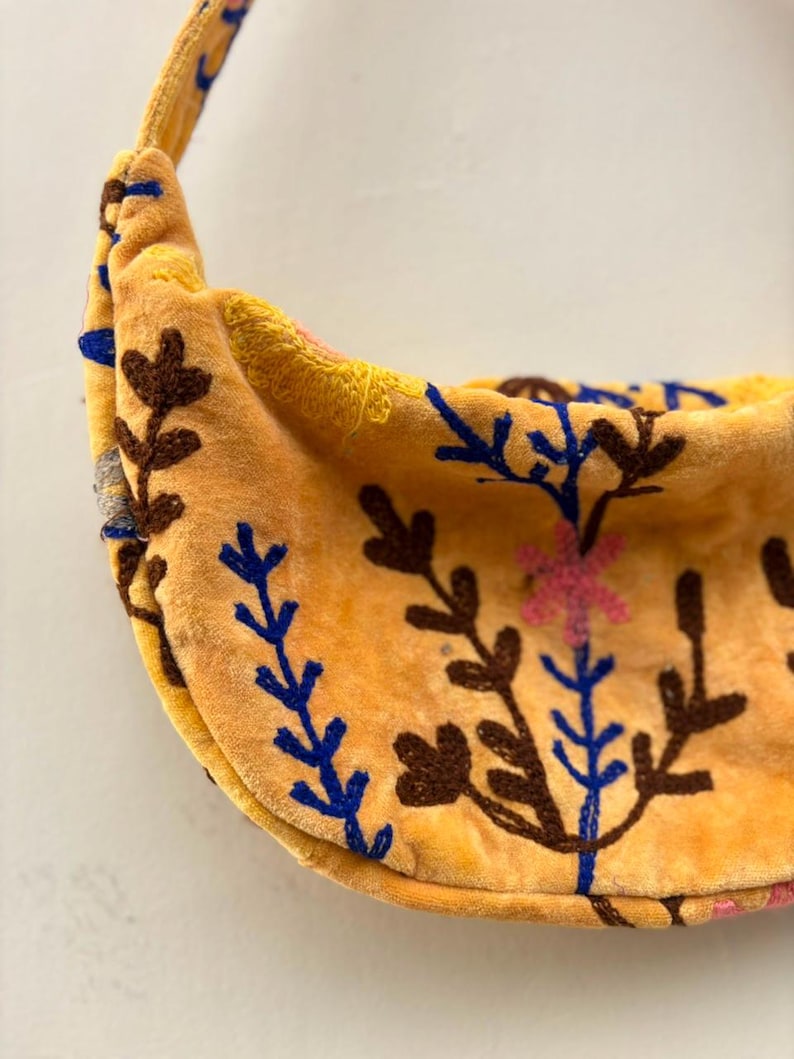 Pu&ograve; includere: Una borsa a tracolla a forma di mezzaluna realizzata in tessuto di velluto giallo. La borsa presenta un intricato ricamo con fili marrone scuro, blu e rosa, che raffigura vari motivi floreali e botanici sulla parte anteriore e sulla tracolla.