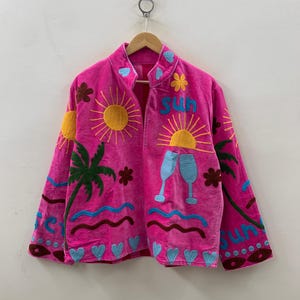 May include: A vibrant pink jacket adorned with embroidered motifs. The design includes a sun, palm trees, flowers, champagne glasses, and the word "SUN". The embroidery is in yellow, blue, brown, and green.