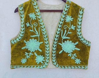 May include: Olive green velvet vest with light blue floral embroidery. The vest is sleeveless and has a cream-colored lining. The embroidery features stylized flowers and vines along the edges and front panels. The vest is hanging on a wooden hanger.