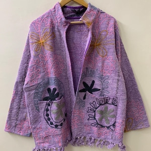 May include: A lavender jacket with embroidered floral designs in pink, yellow, and grey. The jacket has two pockets, a fringe hem, and is hanging on a wooden hanger. The jacket is made of a textured fabric.