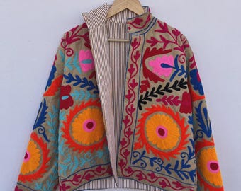 May include: A tan embroidered jacket with colorful floral designs in orange, pink, blue, and red. The jacket has a striped lining and a high collar. The jacket is hanging on a wooden hanger.