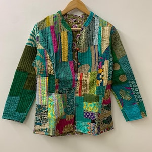 Könnte beinhalten: Patchwork-Jacke mit Stehkragen und langen Ärmeln. Die Jacke hat eine Vielzahl von Stoffen in Grün-, Türkis-, Rosa- und Goldtönen. Sie hat zwei Vordertaschen und wird mit Knöpfen geschlossen. Die Jacke hängt an einem Holzbügel.