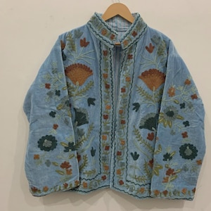 May include: Light blue embroidered jacket with a mandarin collar. The jacket features intricate floral embroidery in shades of brown, green, and teal. The jacket is hanging on a wooden hanger against a neutral background. The jacket is reversible.