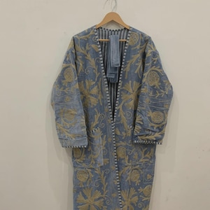 May include: A long, light blue coat with intricate gold floral embroidery. The coat features a V-neck, long sleeves, and two pockets. A matching scarf hangs inside the coat. The coat is hanging on a wooden hanger.