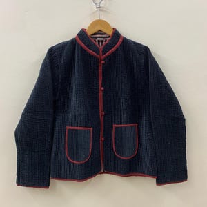 May include: A navy blue corduroy jacket with red trim and two front pockets. The jacket has a mandarin collar and red buttons. The jacket is hanging on a wooden hanger against a plain white wall.
