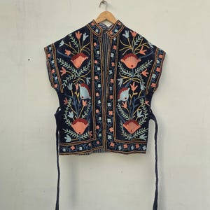 Puede incluir: Chaqueta bordada azul marino con cuello mandarín y mangas cortas. La chaqueta presenta un diseño floral en tonos coral, azul claro y amarillo. Tiene cierres de lazo en la cintura y está colgada en una percha de madera.