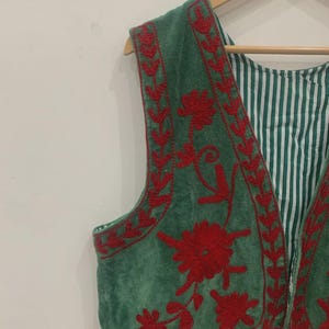 K&ouml;nnte beinhalten: Eine gr&uuml;ne Weste mit roter Blumenstickerei. Die Weste hat eine weiche, Wildleder-&auml;hnliche Textur und ein gestreiftes Futter. Die Stickerei zeigt detaillierte Blumenmuster an den R&auml;ndern und auf der Vorderseite. Die Weste h&auml;ngt an einem Holzb&uuml;gel.