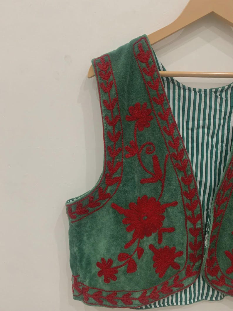 K&ouml;nnte beinhalten: Eine gr&uuml;ne Weste mit roter Blumenstickerei. Die Weste hat eine weiche, Wildleder-&auml;hnliche Textur und ein gestreiftes Futter. Die Stickerei zeigt detaillierte Blumenmuster an den R&auml;ndern und auf der Vorderseite. Die Weste h&auml;ngt an einem Holzb&uuml;gel.