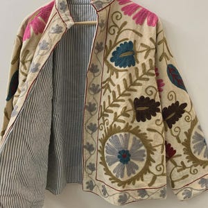 Puede incluir: Chaqueta beige bordada con dise&ntilde;os florales y de hojas en rosa, azul y marr&oacute;n. La chaqueta tiene un forro a rayas y ribetes rojos. La chaqueta est&aacute; colgada en una percha de madera.
